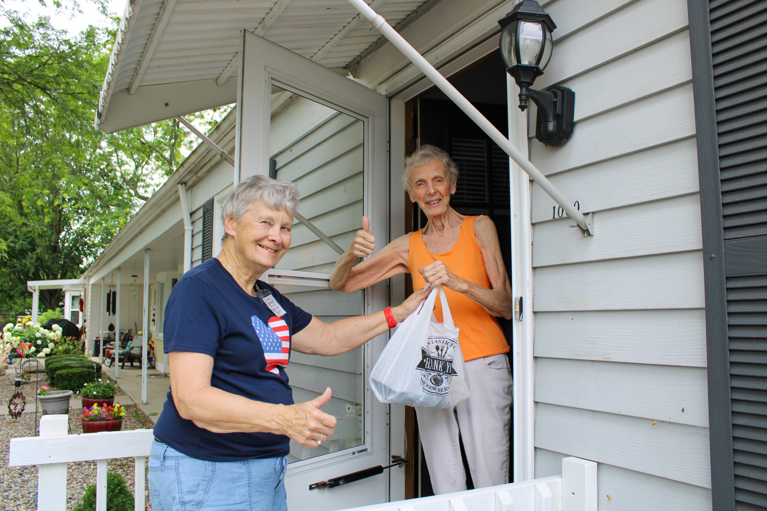 Meals at Home Delivery Image of woman delivering food to another woman, both smiling with thumbs up.