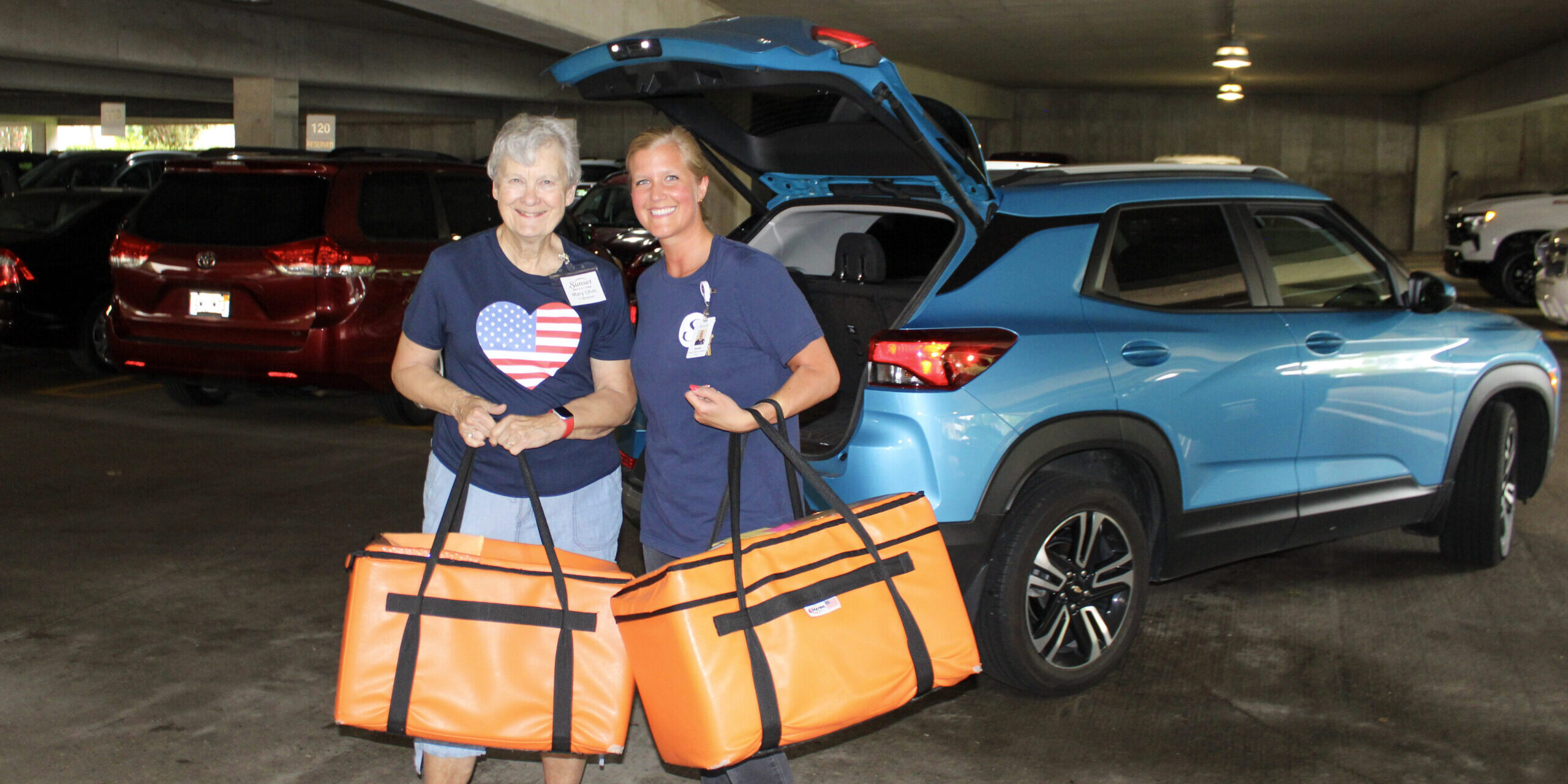 Meals at Home Pickup 2026 Two ladies holding orange bags filled with food for Sunset at Home delivery
