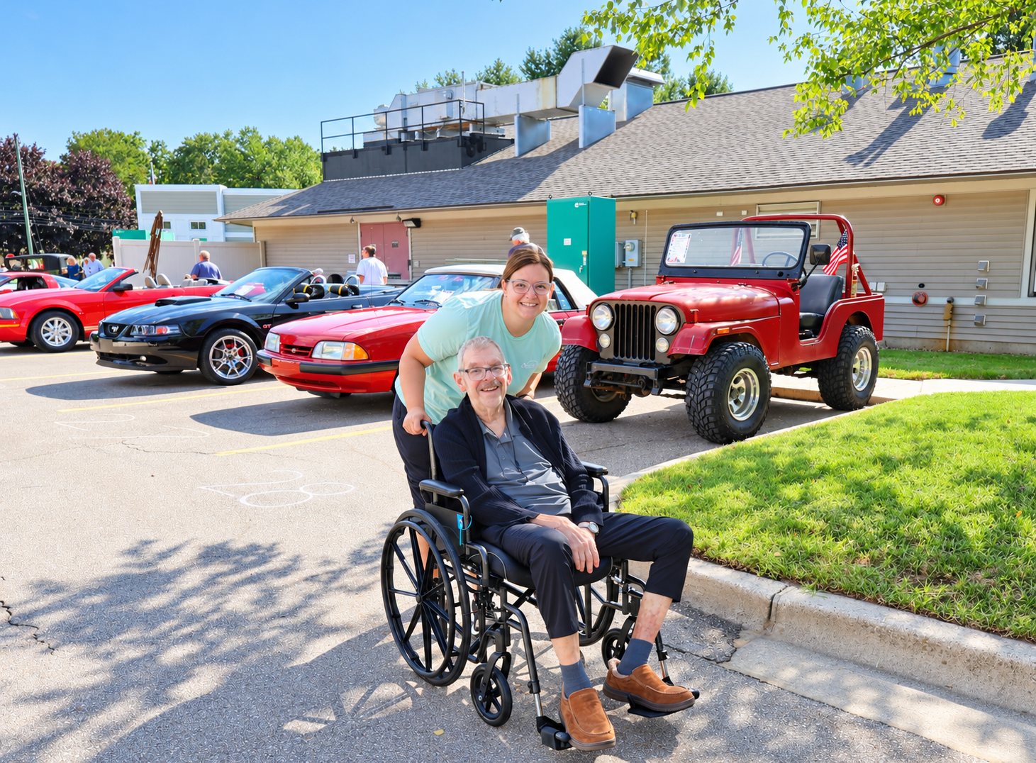 Photo of female staff member and male resident in a wheelchair walking outside during a car show.